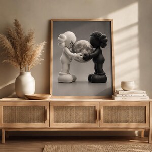 May include: A framed wall art print featuring a white figure and a black figure facing each other, both holding a textured gray sphere between them. The figures have rounded ears with X-shaped eyes and are depicted against a neutral gray background.