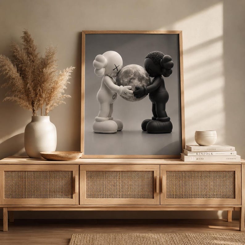 May include: A framed wall art print featuring a white figure and a black figure facing each other, both holding a textured gray sphere between them. The figures have rounded ears with X-shaped eyes and are depicted against a neutral gray background.