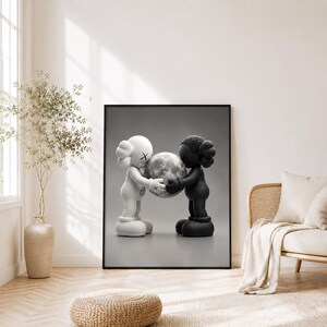 May include: A framed wall art print featuring a black and white illustration of two cartoon-style figures with X-shaped eyes. One figure is white and the other is black, and they are both holding a textured, spherical moon between their hands.