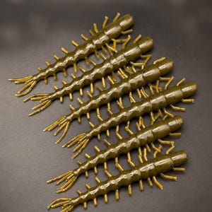 May include: Six soft plastic fishing lures shaped like centipedes. Each lure features a segmented olive green body with small dark speckles and numerous thin, light yellow appendages extending from the sides, ending in a multi-pronged tail.