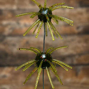 May include: Two olive green, spherical fishing lures with multiple radiating, translucent, flexible appendages. Each lure features a smooth, dark green core embedded with small, reflective orange and black glitter particles throughout the body.