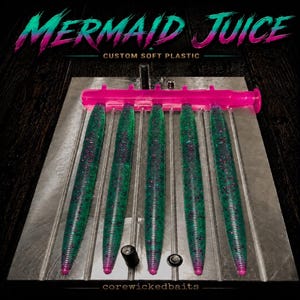 May include: A metal injection mold containing five green and purple glitter soft plastic fishing lures. The top of the mold features the text MERMAID JUICE CUSTOM SOFT PLASTIC, and the bottom includes the text corewickedbaits in small font.