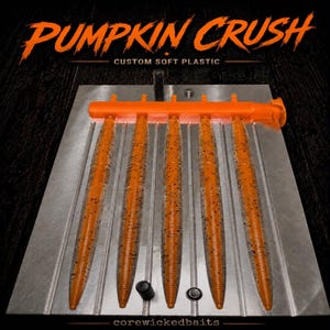 May include: A metal injection mold for fishing lures featuring five parallel cavities filled with orange soft plastic bait with black flecks. The mold has an orange sprue bar at the top and is labeled with the text PUMPKIN CRUSH CUSTOM SOFT PLASTIC.