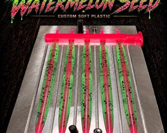 May include: A metal injection mold containing five soft plastic fishing lures. The lures are green with a red center stripe and black speckles. The text WATERMELON SEED CUSTOM SOFT PLASTIC appears at the top, and corewickedbaits is at the bottom.