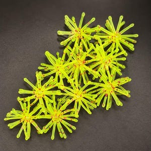 May include: A collection of ten neon yellow, star-shaped fishing lures arranged in a cluster. Each lure features multiple radiating, flexible arms with small, distinct red speckles concentrated near the center point of the star-shaped body.