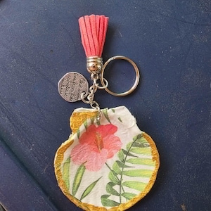 May include: A keyring featuring a scallop shell painted with a pink hibiscus flower and green fern leaves, edged in gold. It includes a coral-coloured suede tassel and a silver-toned metal charm engraved with the text: And every fair from fair sometimes declines.