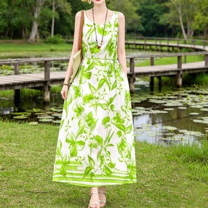 Puede incluir: Un vestido largo sin mangas con fondo blanco y un vibrante estampado floral verde. El vestido presenta un cintur&oacute;n a juego y un dobladillo a rayas. La modelo lleva sandalias plateadas y un collar. El fondo incluye un puente de madera y una masa de agua.