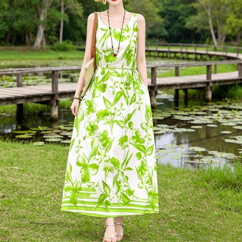 Puede incluir: Un vestido largo sin mangas con fondo blanco y un vibrante estampado floral verde. El vestido presenta un cintur&oacute;n a juego y un dobladillo a rayas. La modelo lleva sandalias plateadas y un collar. El fondo incluye un puente de madera y una masa de agua.