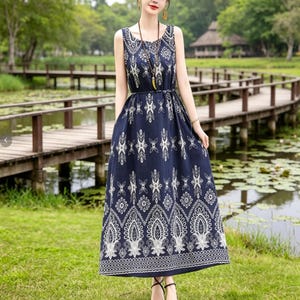 Puede incluir: Un vestido largo sin mangas de color azul marino con un estampado floral blanco. El vestido tiene una cintura ajustada y una falda fluida. La modelo lleva un collar y sandalias. El fondo muestra un puente de madera sobre un estanque.