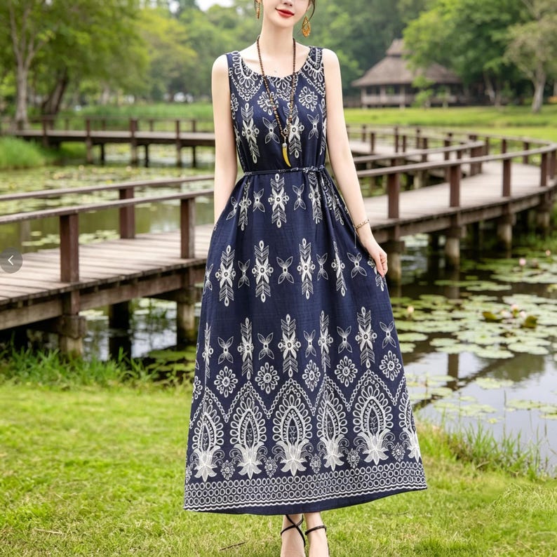 Puede incluir: Un vestido largo sin mangas de color azul marino con un estampado floral blanco. El vestido tiene una cintura ajustada y una falda fluida. La modelo lleva un collar y sandalias. El fondo muestra un puente de madera sobre un estanque.