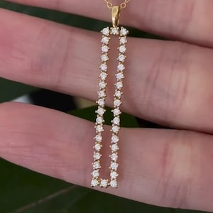 Peut inclure: Un collier en or avec un pendentif rectangulaire long, orné de petites pierres rondes transparentes. Le pendentif est suspendu à une fine chaîne en or. Le texte DiamondsbySVN est imprimé en noir près du haut de la chaîne en or.