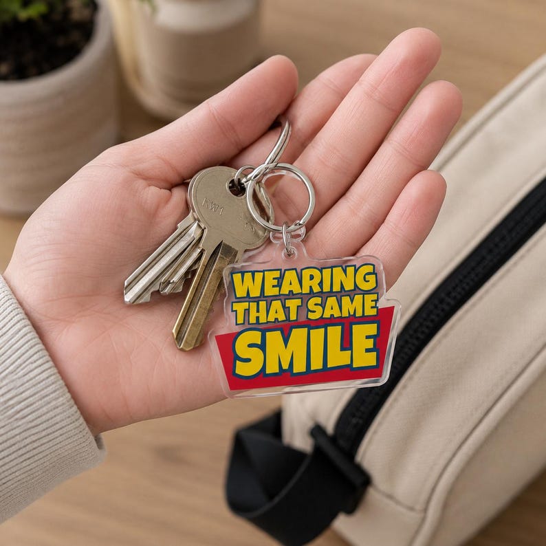 Peut inclure: Un porte-cl&eacute;s en acrylique transparent avec un motif rectangulaire portant le texte jaune WEARING THAT SAME SMILE sur un fond rouge. Le porte-cl&eacute;s est fix&eacute; &agrave; un anneau fendu en m&eacute;tal argent&eacute; qui maintient un jeu de cl&eacute;s en m&eacute;tal argent&eacute;.