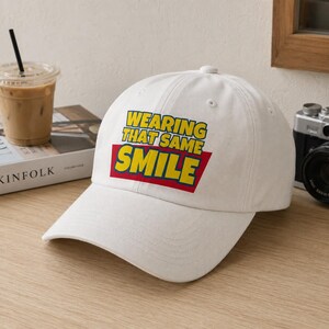 Peut inclure: Une casquette de baseball blanche avec un motif graphique affichant le texte jaune "WEARING THAT SAME SMILE" sur un fond trap&eacute;zo&iuml;dal rouge. La casquette pr&eacute;sente une calotte structur&eacute;e, une visi&egrave;re incurv&eacute;e et un bouton au sommet du d&ocirc;me.