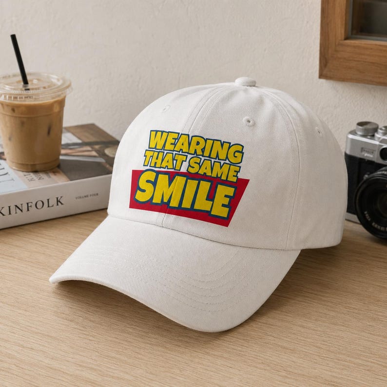 Peut inclure: Une casquette de baseball blanche avec un motif graphique affichant le texte jaune "WEARING THAT SAME SMILE" sur un fond trap&eacute;zo&iuml;dal rouge. La casquette pr&eacute;sente une calotte structur&eacute;e, une visi&egrave;re incurv&eacute;e et un bouton au sommet du d&ocirc;me.