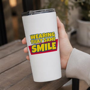 Peut inclure: Un gobelet cylindrique blanc avec un couvercle noir portant le texte "WEARING THAT SAME SMILE" en lettres jaunes sur un fond rouge. Le motif est centr&eacute; sur le devant du r&eacute;cipient isotherme, qui pr&eacute;sente une finition lisse et uniforme.