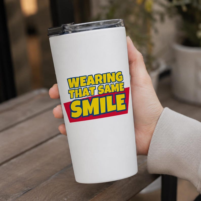 Peut inclure: Un gobelet cylindrique blanc avec un couvercle noir portant le texte "WEARING THAT SAME SMILE" en lettres jaunes sur un fond rouge. Le motif est centr&eacute; sur le devant du r&eacute;cipient isotherme, qui pr&eacute;sente une finition lisse et uniforme.