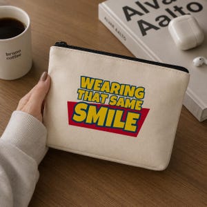 Peut inclure: Une pochette rectangulaire en toile blanc cass&eacute; avec le texte "WEARING THAT SAME SMILE" imprim&eacute; en lettres jaunes grasses avec un contour bleu, sur un fond trap&eacute;zo&iuml;dal rouge. La pochette poss&egrave;de une fermeture &eacute;clair noire sur le dessus.