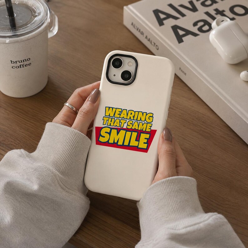 Peut inclure: Une coque de smartphone couleur cr&egrave;me arborant un motif graphique avec le texte "WEARING THAT SAME SMILE" en lettres jaunes sur fond rouge. La coque pr&eacute;sente une d&eacute;coupe noire pour l'objectif de l'appareil photo et est tenue en main.