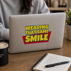 Peut inclure: Un autocollant rectangulaire appliqu&eacute; sur le couvercle d'un ordinateur portable argent&eacute;. L'autocollant pr&eacute;sente le texte WEARING THAT SAME SMILE en lettres jaunes grasses avec un contour bleu, sur un fond trap&eacute;zo&iuml;dal rouge.