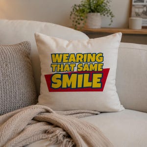 Peut inclure: Un coussin carr&eacute; blanc cass&eacute; avec le texte WEARING THAT SAME SMILE en lettres jaunes grasses. Le mot SMILE est plac&eacute; sur un fond trap&eacute;zo&iuml;dal rouge uni. Le coussin pr&eacute;sente une texture de tissu lisse et unie ainsi qu'un design simple et &eacute;pur&eacute;.