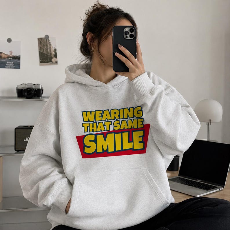 Peut inclure: Un sweat-shirt &agrave; capuche gris clair avec une impression graphique sur la poitrine. Le texte WEARING THAT SAME SMILE est &eacute;crit en lettres jaunes sur un fond rectangulaire rouge. Le sweat poss&egrave;de des manches longues et une poche kangourou.