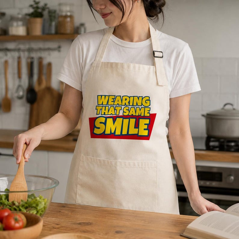 Peut inclure: Un tablier de cuisine beige avec le texte WEARING THAT SAME SMILE en lettres jaunes grasses bord&eacute;es de bleu, sur un fond rectangulaire rouge. Le tablier comprend une poche avant et une sangle de cou r&eacute;glable avec une boucle noire.