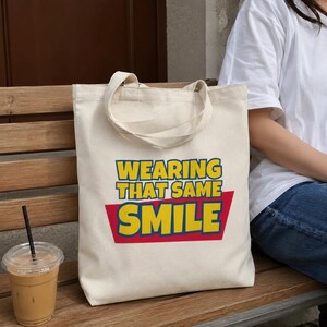 Peut inclure: Un sac fourre-tout en toile de couleur cr&egrave;me avec le texte WEARING THAT SAME SMILE imprim&eacute; en lettres jaunes grasses sur un fond rouge. Le sac poss&egrave;de deux anses en tissu assorties et une forme rectangulaire en mat&eacute;riau tiss&eacute; durable.