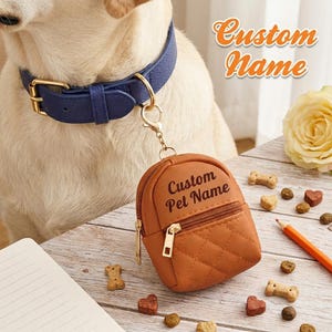 Puede incluir: Un collar de perro de cuero azul con hebilla dorada unido a una pequeña bolsa marrón acolchada con forma de mochila. La bolsa cuenta con una cremallera dorada y el texto Custom Pet Name impreso en negro para guardar accesorios.