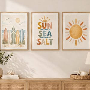 Peut inclure: Un ensemble de trois affiches encadrées. L'affiche de gauche montre cinq planches de surf sur une plage de sable avec un soleil et des oiseaux. Celle du centre porte le texte SUN SEA SALT en lettres colorées. Celle de droite montre un grand soleil.
