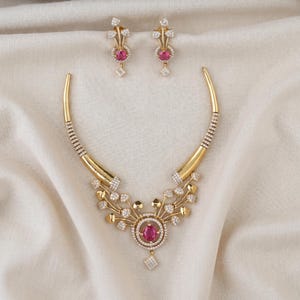 May include: A gold-toned necklace and matching earring set featuring a central circular pendant with a round pink stone, surrounded by small white stones. The necklace has a rigid curved design with leaf-like accents and white stone detailing.