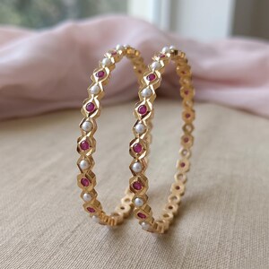Może przedstawiać: Dwie złote bransoletki typu bangle z naprzemiennym wzorem okrągłych białych pereł i okrągłych czerwonych kamieni szlachetnych. Metalowa obręcz ma fasetowany, geometryczny wz&oacute;r, kt&oacute;ry obramowuje każdy kamień, tworząc powtarzającą się sekwencję element&oacute;w.