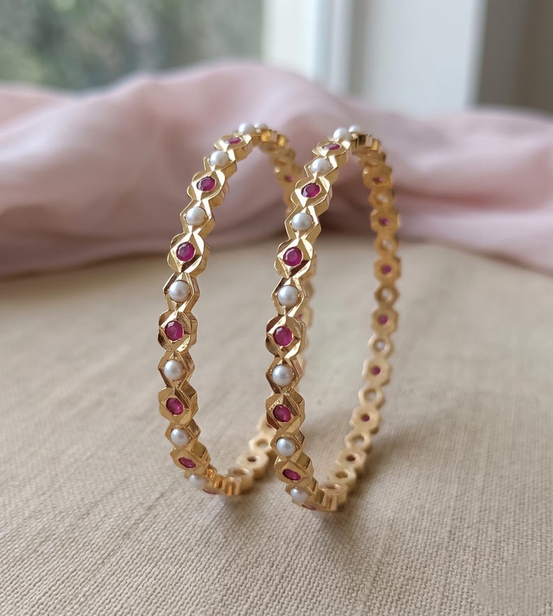 Może przedstawiać: Dwie złote bransoletki typu bangle z naprzemiennym wzorem okrągłych białych pereł i okrągłych czerwonych kamieni szlachetnych. Metalowa obręcz ma fasetowany, geometryczny wz&oacute;r, kt&oacute;ry obramowuje każdy kamień, tworząc powtarzającą się sekwencję element&oacute;w.