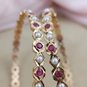 Może przedstawiać: Dwie złote bransoletki typu bangle z naprzemiennym wzorem okrągłych białych pereł i kolistych czerwonych rubin&oacute;w osadzonych w metalowych oprawach w kształcie romb&oacute;w. Złoty metal ma teksturowane, fasetowane wykończenie na krawędziach opraw.