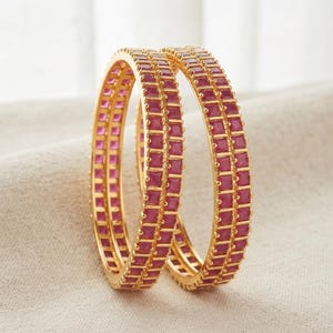 Może przedstawiać: Dwie złote bransoletki typu bangle z podwójnym rzędem kwadratowych, intensywnie różowych kamieni osadzonych w ciągłym kanale. Metaliczne złote wykończenie kontrastuje z głębokim różem kamieni, które ułożone są w jednolity, powtarzalny wzór.
