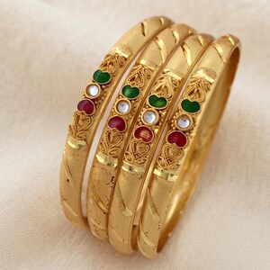Op de afbeelding: Een set van vier goudkleurige armbanden met een matte afwerking en diagonale gegraveerde lijnen. Elke armband is versierd met een centraal decoratief element met een groen geëmailleerd blad, een witte steen en een rood hartvormig accent.