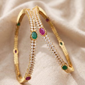 May include: Two gold-toned bangles featuring a textured metal band encrusted with small clear white stones. Each bangle is accented with alternating oval-shaped green and pink gemstones set at intervals along the circular frame of the jewellery.