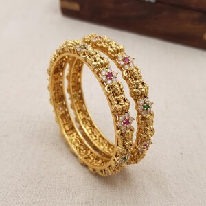 Könnte beinhalten: Zwei goldfarbene Armreifen mit detaillierten, geprägten Gottheiten, die sich mit Blumenmotiven abwechseln. Jede Blume ist mit einem zentralen roten oder grünen Stein besetzt, der von kleinen klaren weißen Steinen umgeben ist, was eine strukturierte Oberfläche ergibt.