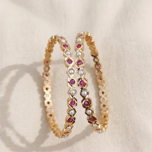 Może przedstawiać: Dwie złote bransoletki typu bangle z naprzemiennym wzorem okrągłych czerwonych kamieni szlachetnych i małych białych pereł osadzonych w geometrycznej, fasetowanej metalowej ramie. Okrągłe bransoletki mają polerowane wykończenie i sp&oacute;jny wz&oacute;r.