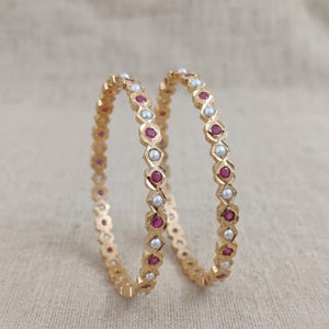 Może przedstawiać: Dwie złote bransoletki typu bangle z powtarzającym się geometrycznym wzorem. Każda bransoletka jest ozdobiona naprzemiennie okrągłymi czerwonymi kamieniami szlachetnymi i małymi białymi perłami. Metalowa powierzchnia ma fasetowaną teksturę, tworząc szereg połączonych ogniw w kształcie romb&oacute;w, kt&oacute;re utrzymują kamienie na całym obwodzie okrągłej biżuterii.