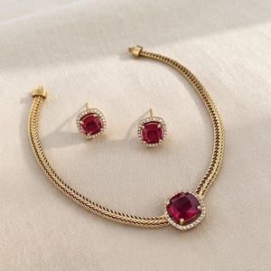 Op de afbeelding: Een goudkleurige ketting en bijpassende oorknopjes. De ketting heeft een gevlochten schakel met een centrale vierkante dieprode edelsteen omringd door kleine witte steentjes. Elke oorbel heeft een bijpassende vierkante rode steen met een rand.