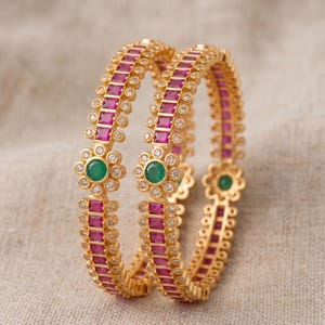 May include: Two gold bangles featuring a repeating pattern of rectangular magenta stones and small clear white stones. Each bangle is accented with a central flower-shaped motif containing a round green stone surrounded by small clear white stones.