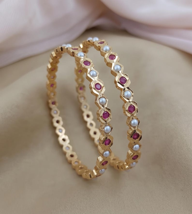 Może przedstawiać: Dwie złote bransoletki typu bangle z naprzemiennym wzorem okrągłych białych pereł i kolistych czerwonych kamieni szlachetnych. Metalowa obręcz ma geometryczny, fasetowany wz&oacute;r, kt&oacute;ry otacza każdy kamień, tworząc ciągłą, powtarzalną strukturę.