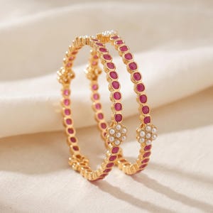 Puede incluir: Dos brazaletes dorados con una hilera continua de piedras redondas de color rosa intenso engastadas en metal. Cada brazalete está acentuado con un pequeño motivo floral de cinco pétalos compuesto por diminutas perlas blancas.
