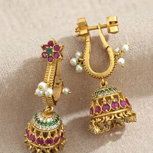 May include: A pair of gold-toned jhumka earrings featuring a flower-shaped stud with a central green stone surrounded by pink petals. The bell-shaped drop is adorned with pink and green stones, clear crystals, and small white pearl-like beads.