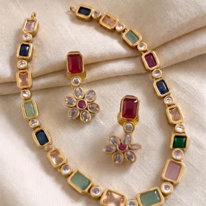 May include: A gold-toned necklace and matching pair of earrings featuring rectangular multi-coloured gemstones and small round clear stones. The necklace consists of alternating rectangular and round links, while each earring has a rectangular stone above a flower-shaped cluster of clear stones.