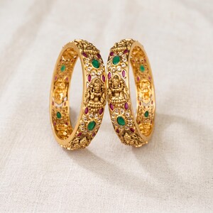 May include: Two gold bangles featuring intricate embossed deity figures, accented with oval-shaped green stones, small red stones, and clear white stones. The metal surface has a textured, matte finish with detailed carvings throughout the circular bands.