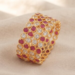 Peut inclure: Une bague dorée présentant un design large composé de quatre rangées interconnectées. Chaque rangée est ornée de pierres circulaires transparentes alternant avec des pierres circulaires rose-rouge, le tout serti dans un métal doré poli.
