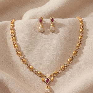 Puede incluir: Un conjunto de collar y pendientes en tono dorado con acabado mate. El collar presenta un patrón de hojas repetitivo con pequeñas piedras claras y un colgante central con una piedra roja talla marquesa y piedras claras en forma de lágrima.