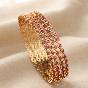Può includere: Un set di tre bracciali rigidi color oro caratterizzati da un design metallico ondulato e sinuoso. Ogni bracciale è impreziosito da una serie di piccole pietre rotonde di colore rosa acceso, incastonate a intervalli regolari lungo la fascia metallica.