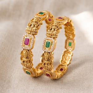 Peut inclure: Deux bracelets rigides dorés ornés d'une alternance de motifs rectangulaires et de figures divines en relief. Les motifs rectangulaires contiennent une pierre vert émeraude ou rouge rubis, entourée d'un bord de petits cristaux blancs.